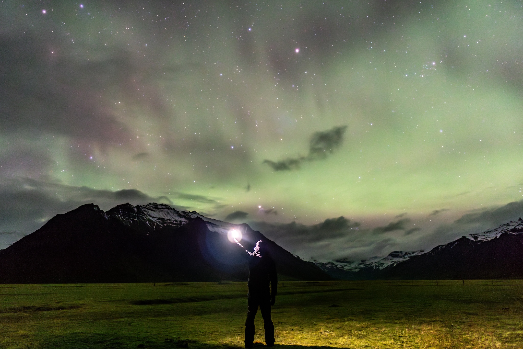 Una persona de espaldas levanta una luz hacia una aurora boreal verde y rosa sobre unas montañas. La escena está iluminada por la luz de la aurora y de la linterna.